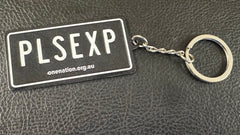 Black “PLS EXP” licence plate keyring on a dark background